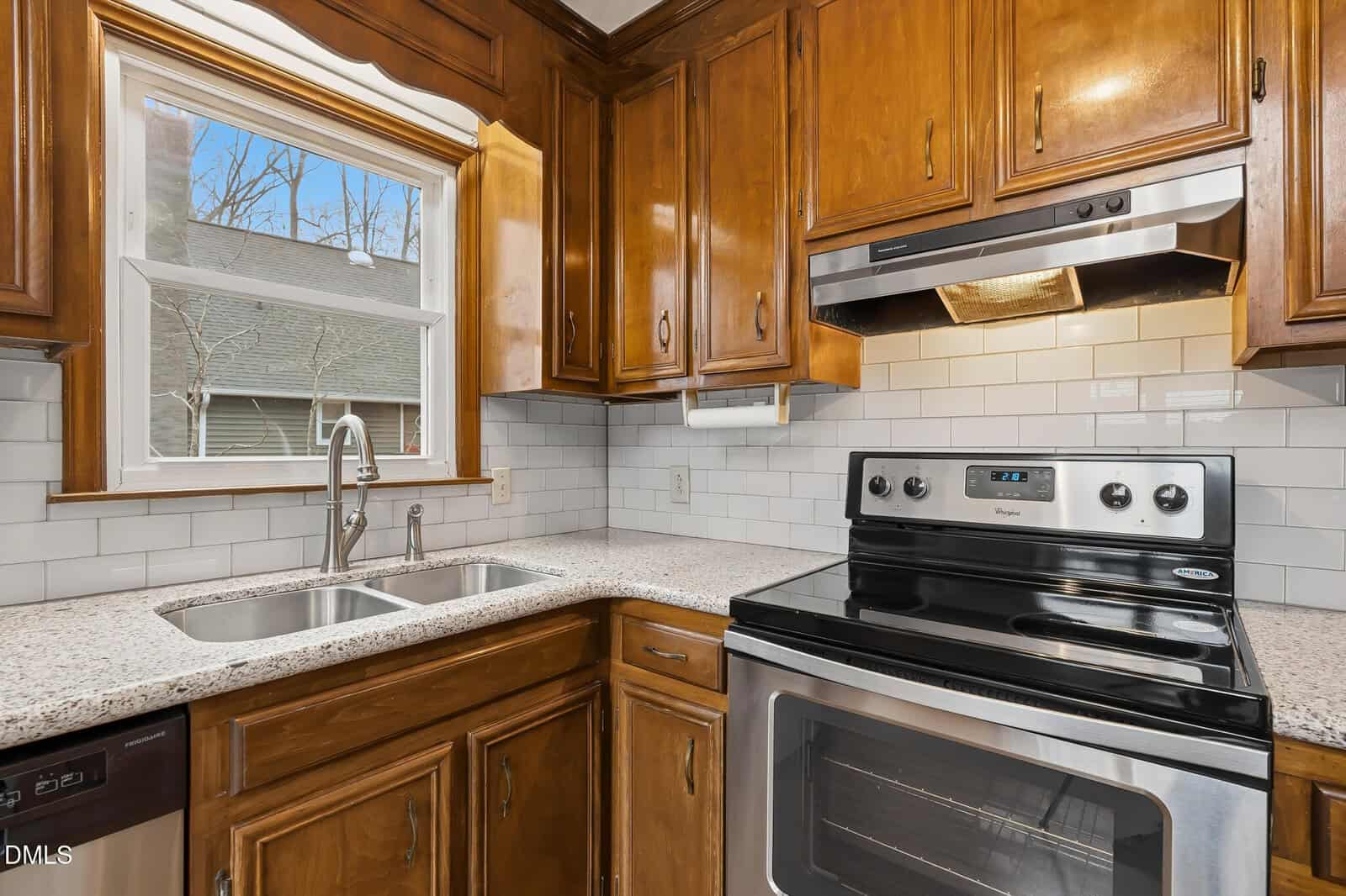 A kitchen with wooden cabinets, a double sink under a window, a stainless steel electric stove and oven, white subway tile backsplash, and granite countertops. Trees and a house are visible through the window.