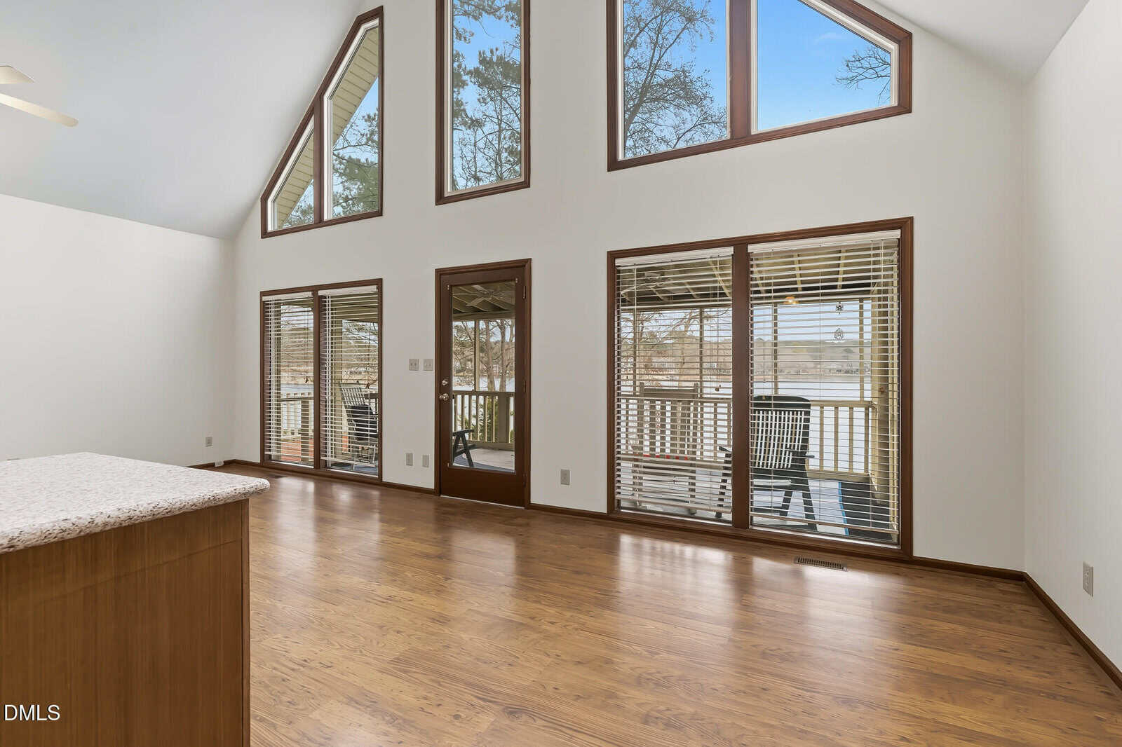 A spacious, empty room with wood flooring, large windows, and glass doors leading to a deck outside. The walls are white, and the ceiling is vaulted. Natural light fills the room through the windows.