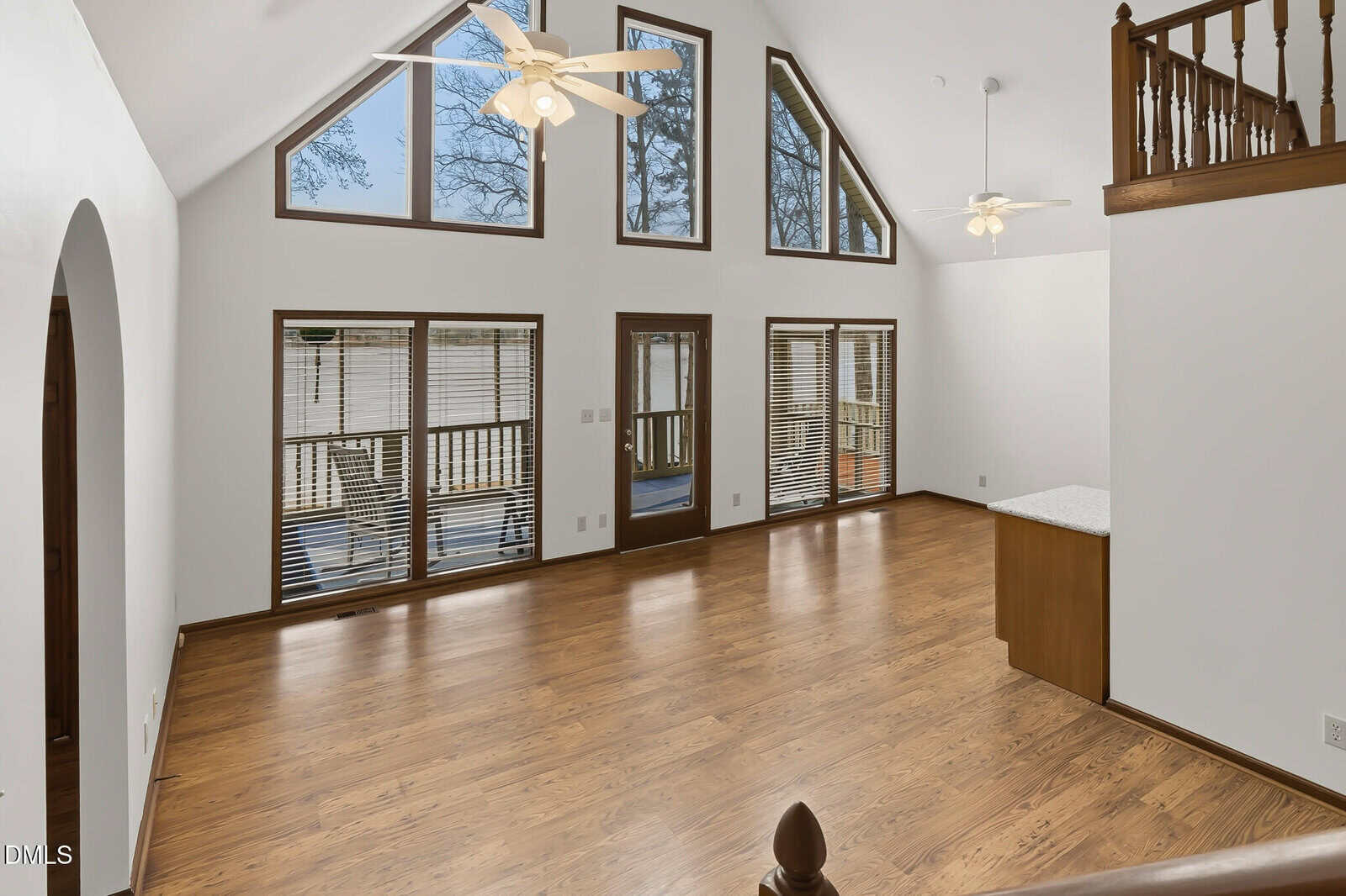 Spacious living area with high vaulted ceiling, large triangular and rectangular windows, ceiling fans, wood flooring, and an open layout leading to a kitchen counter with white surface. Balcony railing visible on upper right.