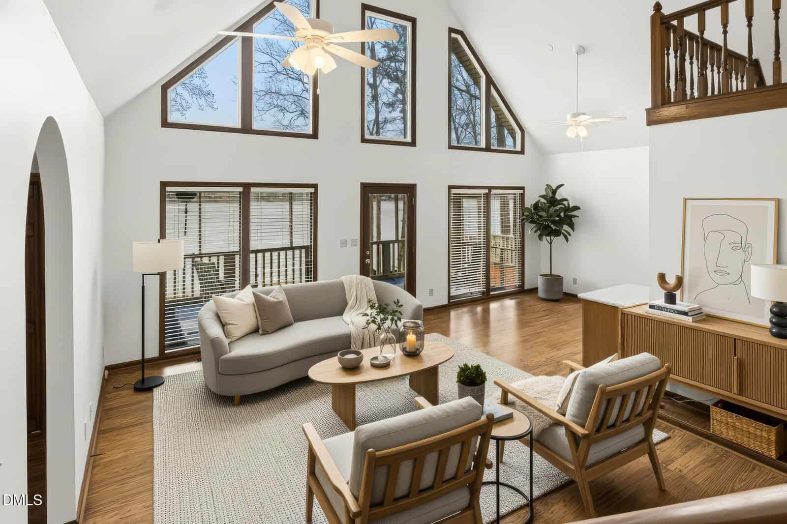 A living room with high vaulted ceilings, large triangular and rectangular windows, two ceiling fans, a gray sofa, two armchairs, a round coffee table, a sideboard, houseplants, and modern decor.