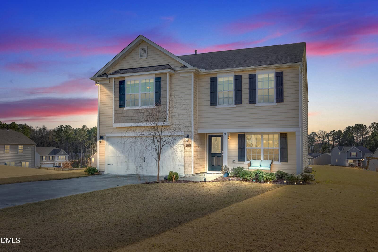 Two-story beige house with dark shutters at 2802 W Brookwood Court, Creedmoor, featuring an attached double garage and small front porch. Exterior lights glow as the home sits on a large grassy lot at dusk under a colorful sky.