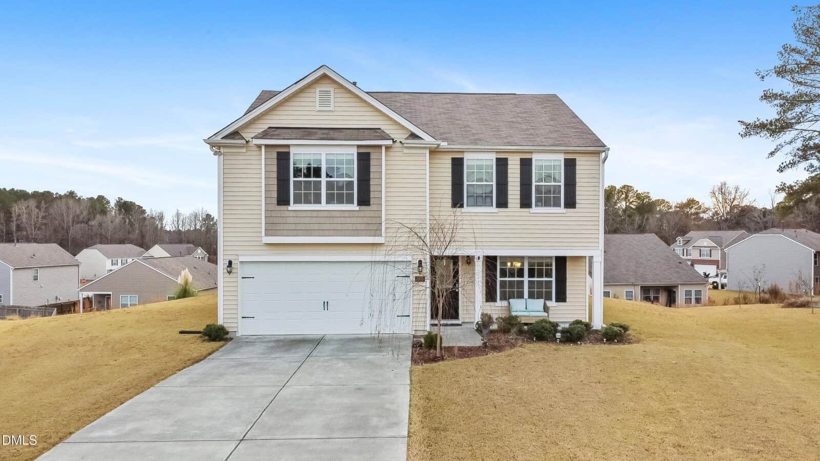 Two-story suburban house with beige siding, white trim, and black shutters. Attached two-car garage, concrete driveway, small front porch, and front yard with minimal landscaping. Neighboring houses and trees in the background.