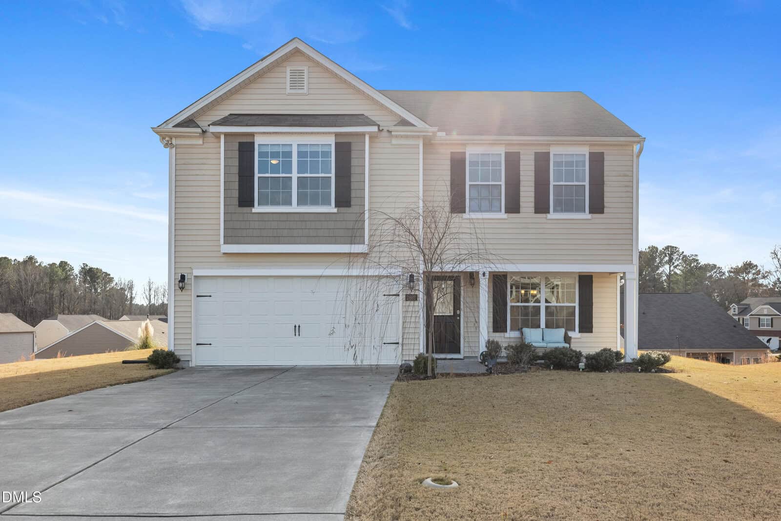 Two-story beige house with white trim, black shutters, and a double garage. A bare tree and small shrubs are in the front yard. The house sits on a sloped lawn with a concrete driveway under a clear blue sky.