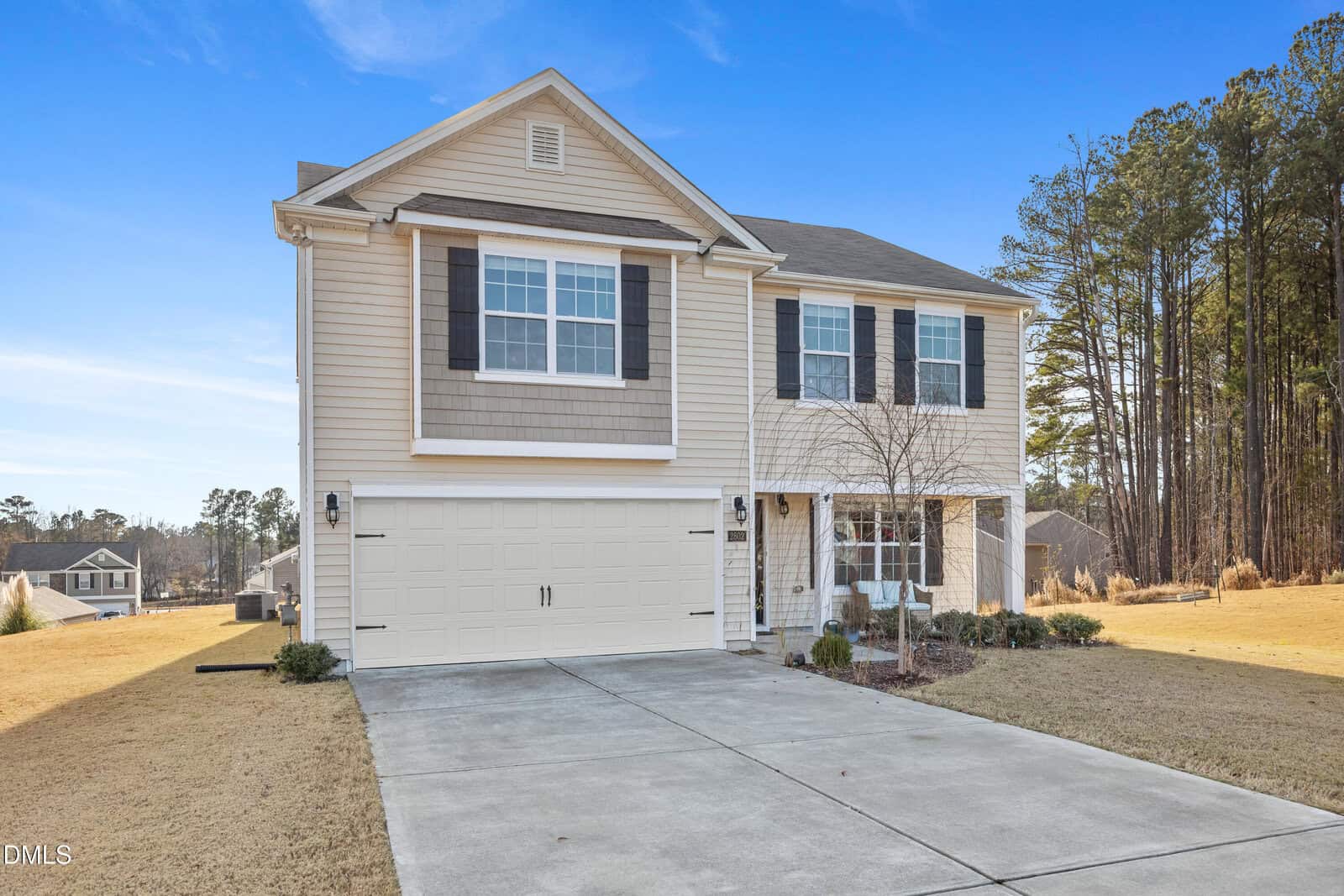 Two-story suburban house with beige siding, white trim, and black shutters. A two-car garage is attached, and a concrete driveway leads up to the house. The yard is grassy with a few small shrubs and trees.