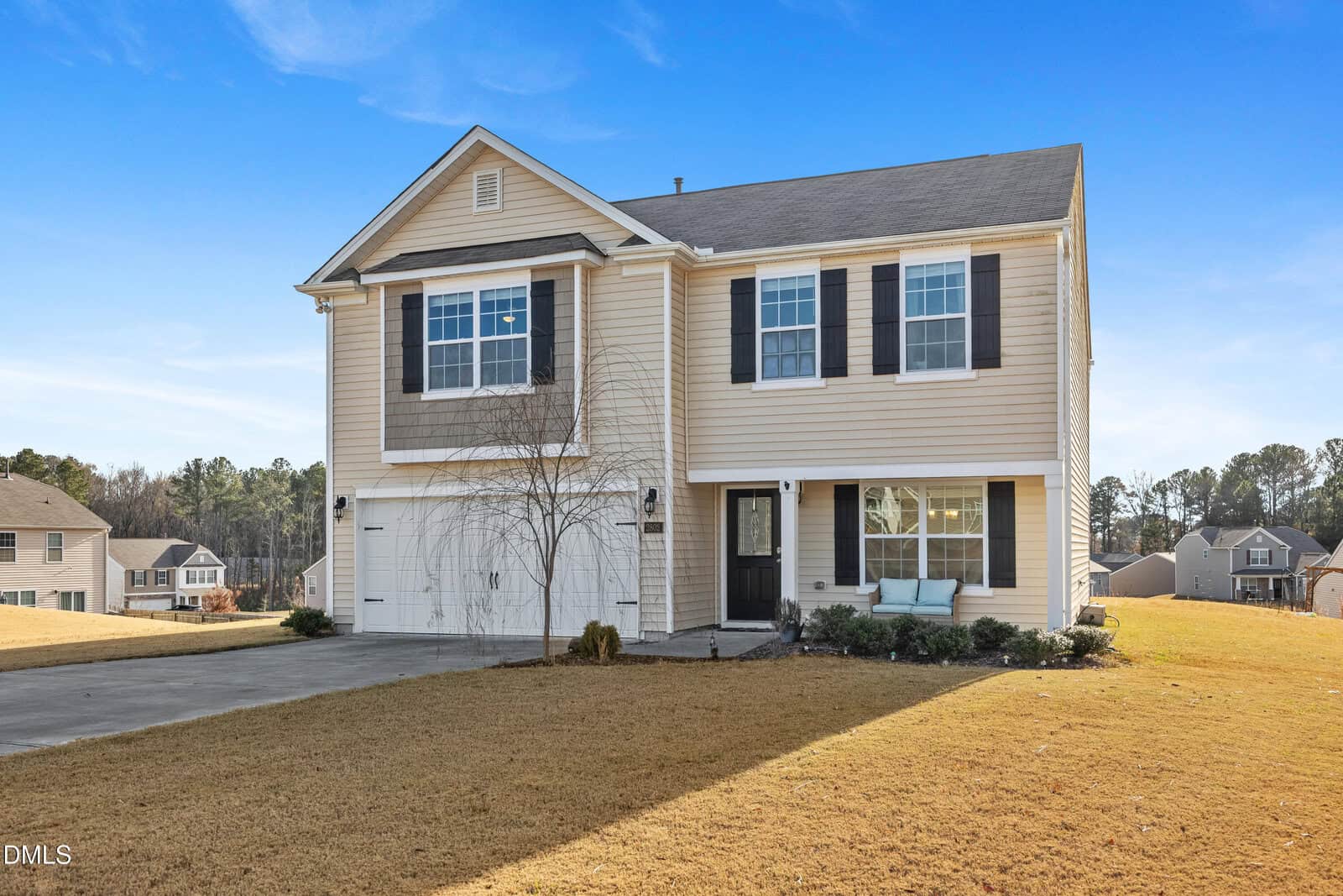 Two-story beige house with black shutters, attached two-car garage, and a small front porch with seating. The house is surrounded by a dry, yellowed lawn and has a clear blue sky in the background.