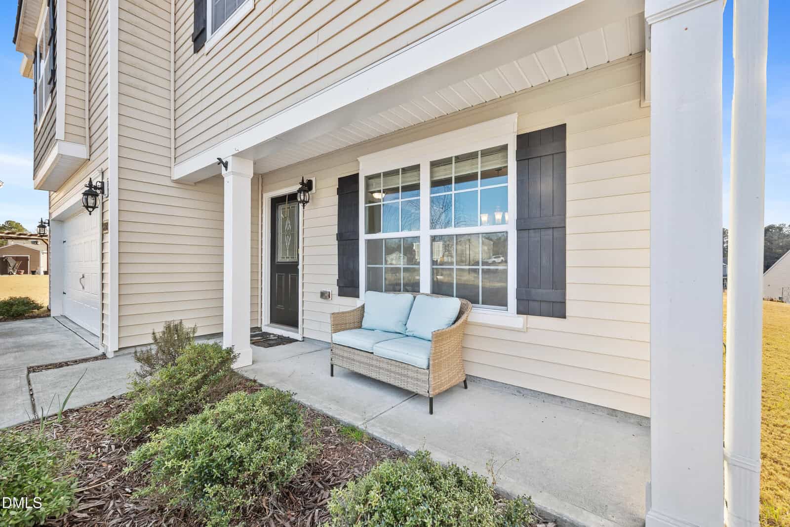 A beige house exterior with white trim features a black front door, a large window with shutters, and a small porch with a wicker loveseat and blue cushions. Shrubs line the edge of the concrete walkway.