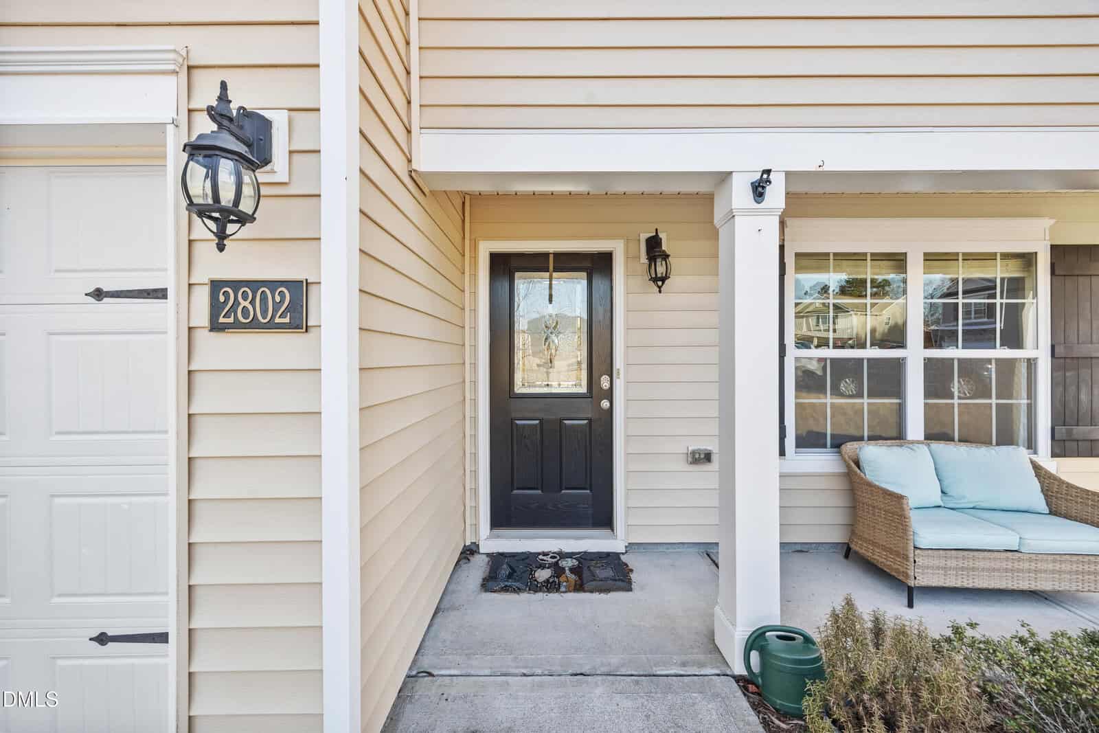 Front porch view of a beige house with black door, wall-mounted lanterns, house number 2802, window with seating area, and a green garden hose on the ground.