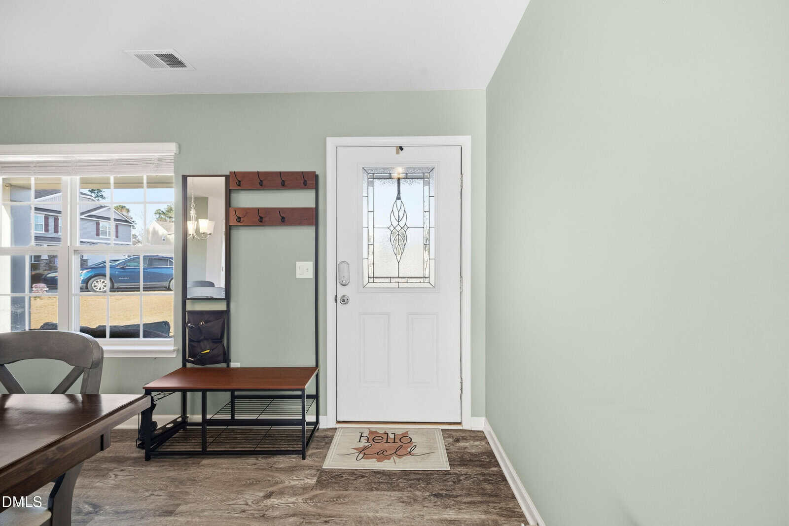 A white front door with a decorative glass panel is centered in a light green wall. A coat rack with a bench is to the left, and a doormat that reads "hello fall" is in front of the door. A window shows a car outside.