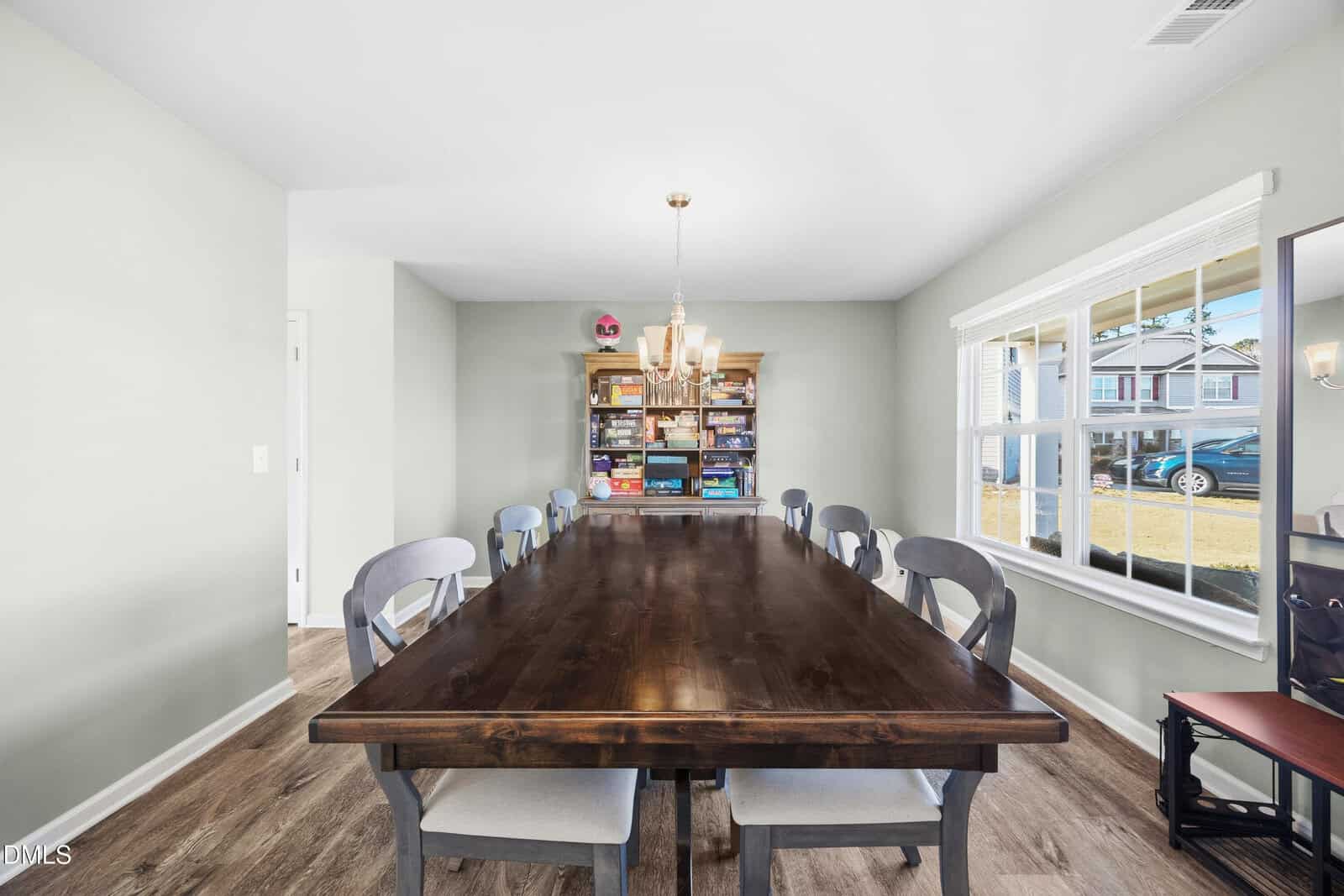 A dining room with a long wooden table surrounded by eight gray chairs. A chandelier hangs above the table, and a bookshelf filled with board games stands against the far wall. Sunlight streams through a large window.