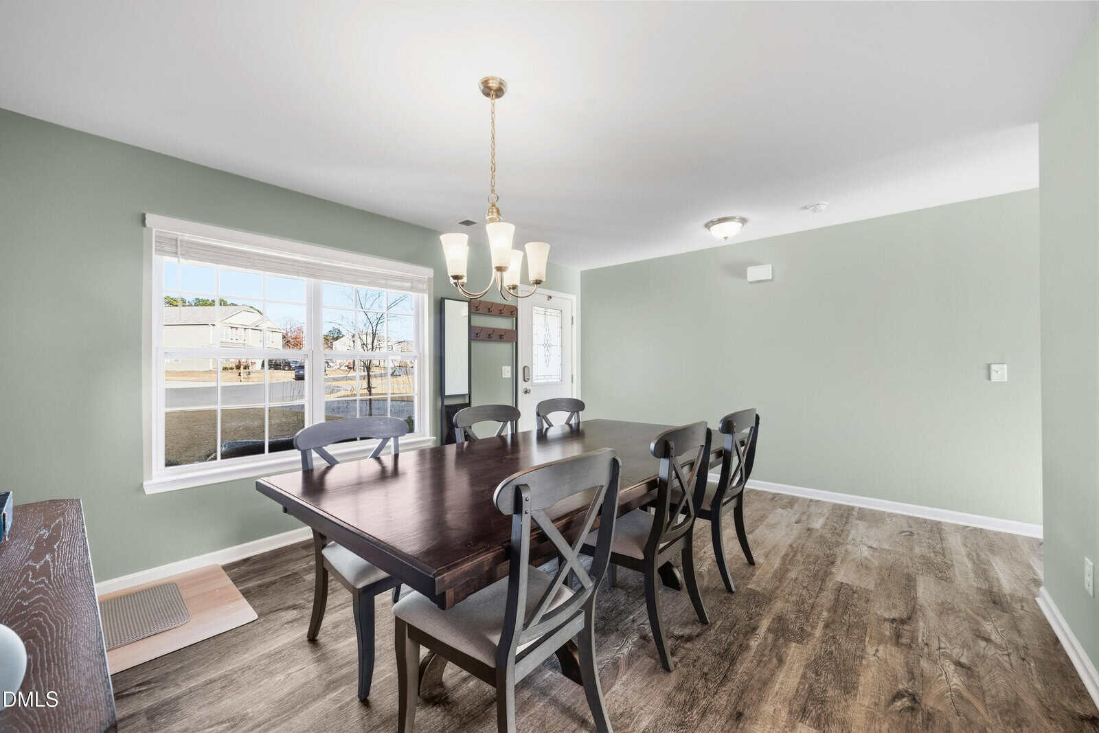 Dining room with a dark wood table and eight chairs, a hanging light fixture above, light green walls, wood flooring, a large window on the left, and a white door at the back of the room.