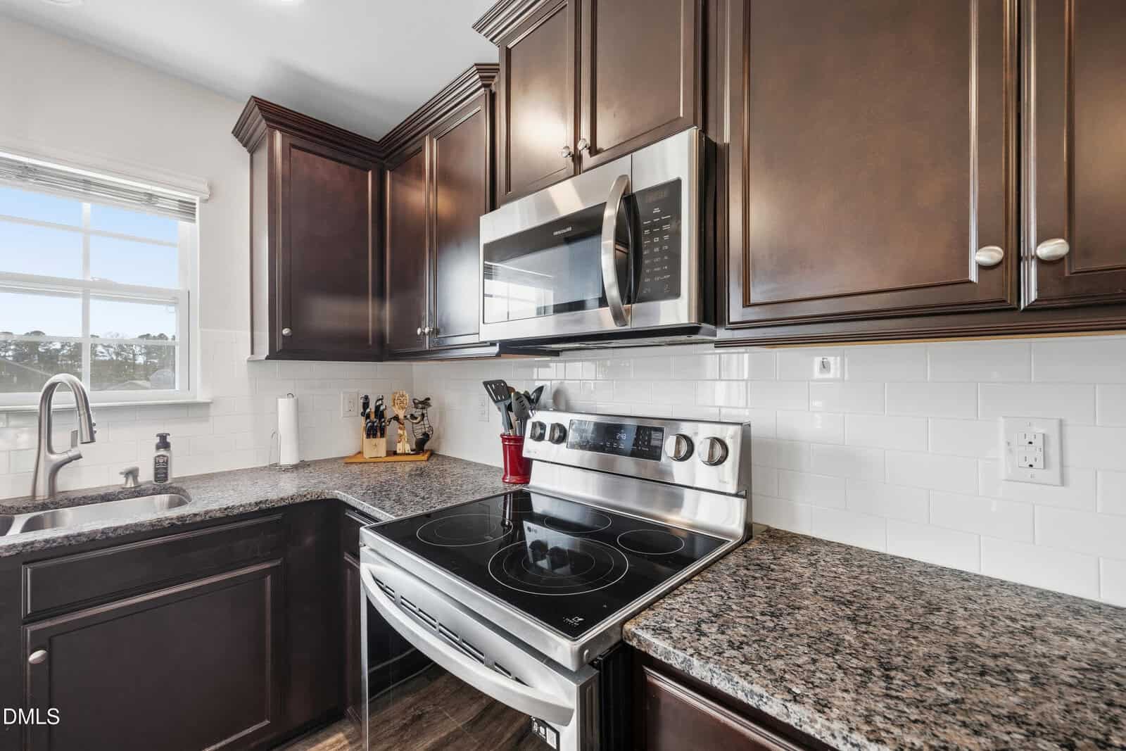 Modern kitchen with dark wood cabinets, granite countertops, stainless steel stove and microwave, a window above the sink, and white subway tile backsplash. Knife block and cooking utensils are on the counter.