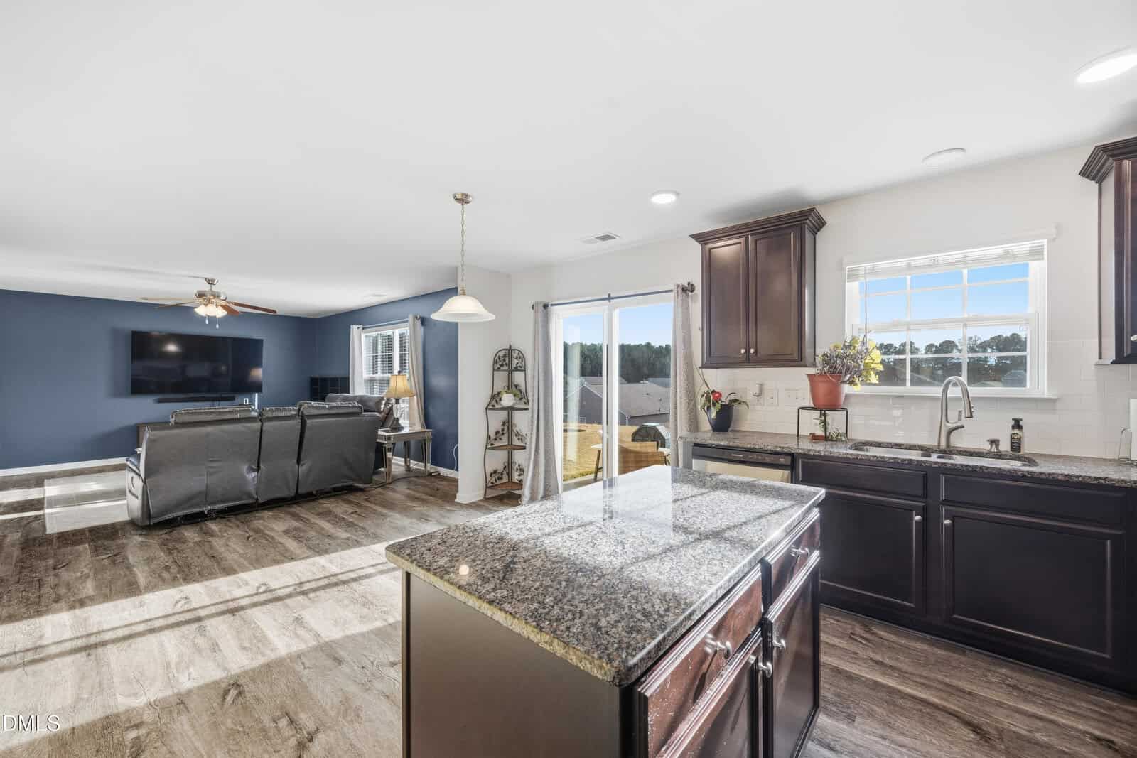 Modern open-concept kitchen with dark cabinets, granite island, and stainless steel appliances, overlooking a living room with recliners, TV, and large windows letting in natural light.