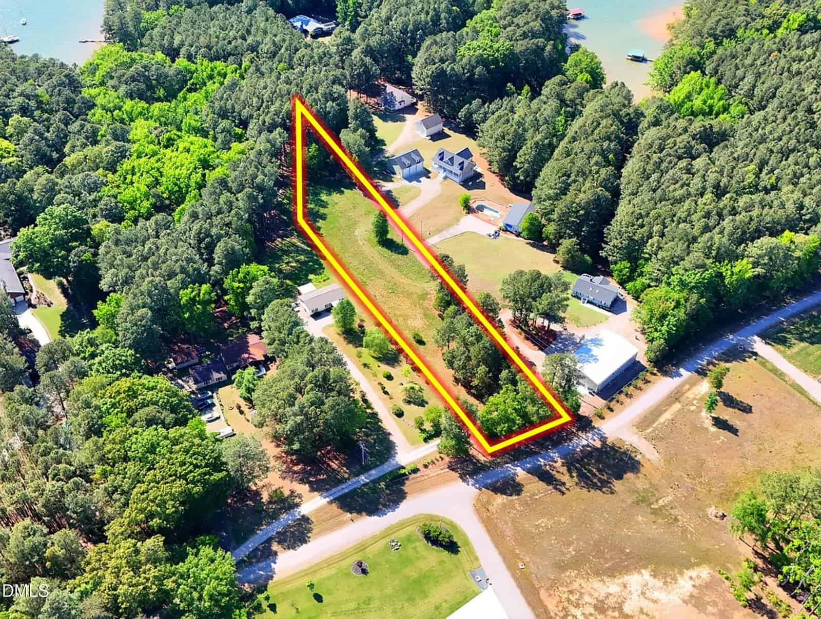 Aerial view of a triangular plot of land outlined in orange, surrounded by trees and neighboring houses, near a road and close to a lake.