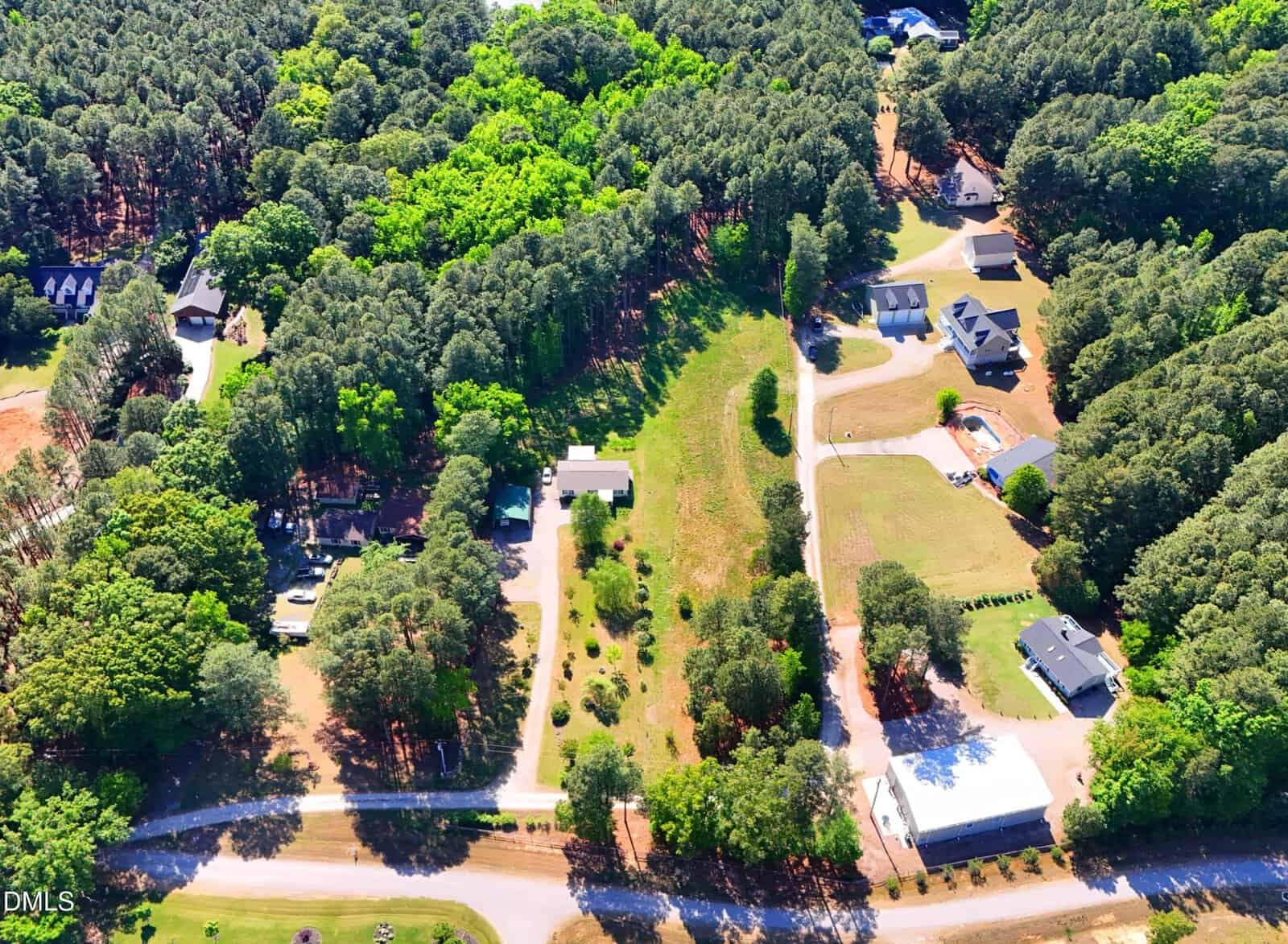 Aerial view of a rural neighborhood with houses, driveways, trees, and grassy areas, surrounded by dense forest and intersected by a road.