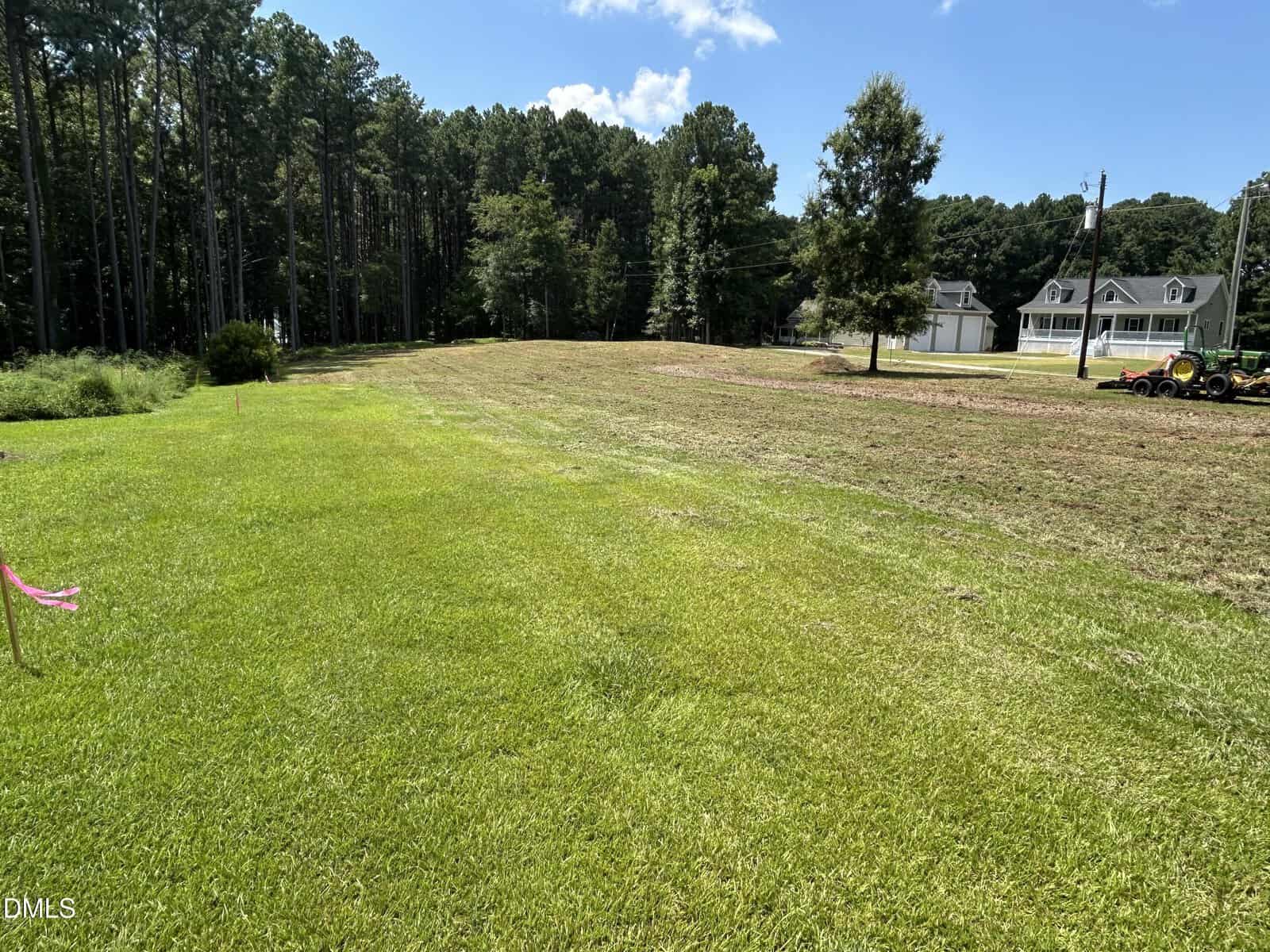 A large grassy lot with trees in the background, a mowed lawn, and a house with cars on the right side under a blue sky with a few clouds.