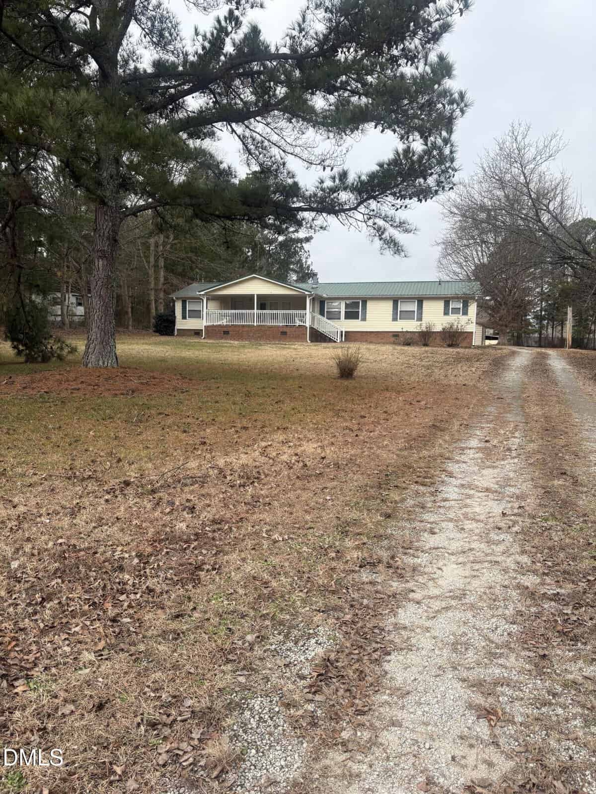 A single-story beige house with a green roof sits at the end of a gravel driveway on 1880 Long Mill Road in Youngsville, surrounded by brown grass, bare trees, and pine trees on a cloudy day.