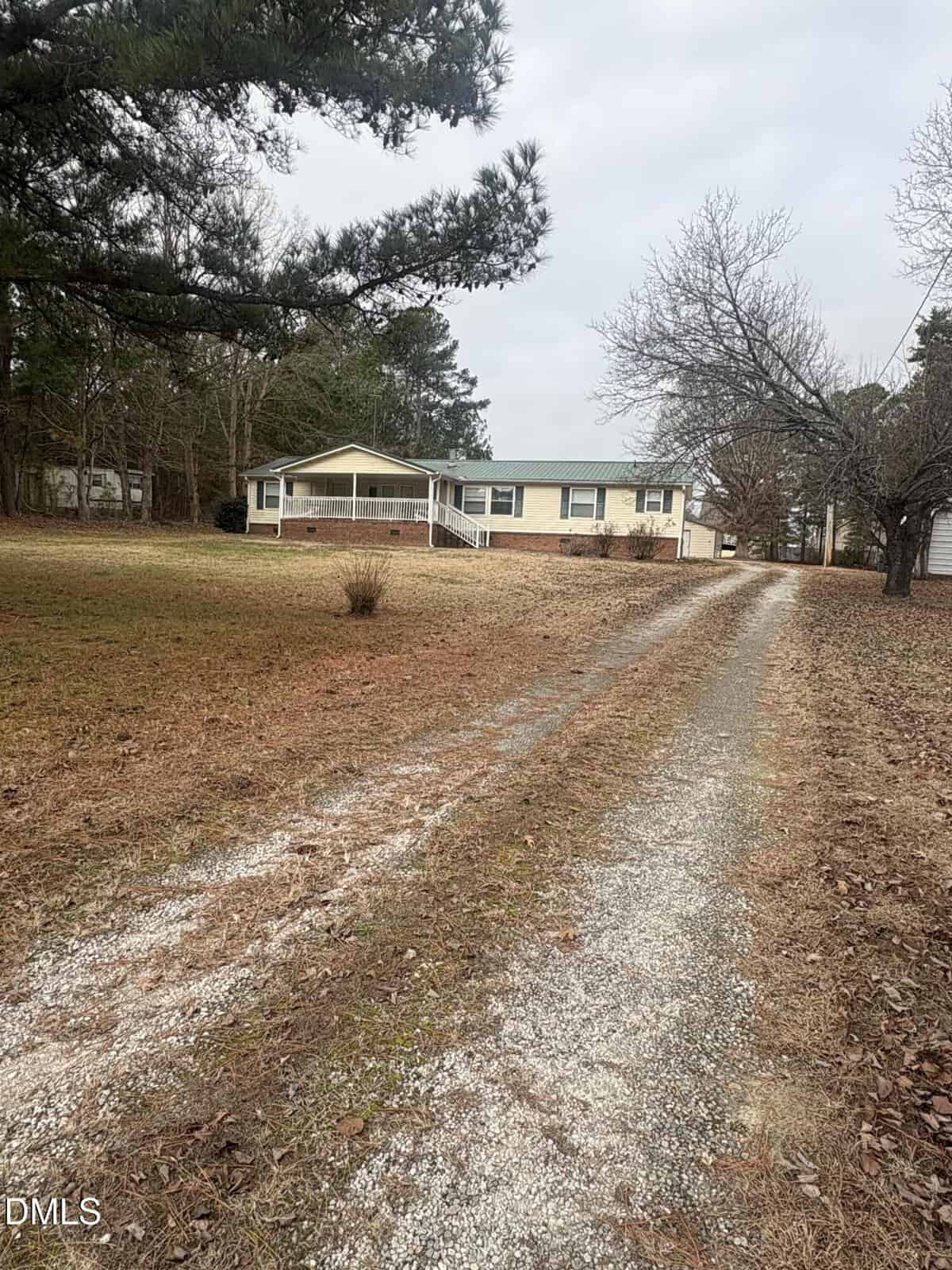 A long gravel driveway leads to a single-story beige house with a green roof and brick foundation. The house is surrounded by bare trees and brown grass under an overcast sky.