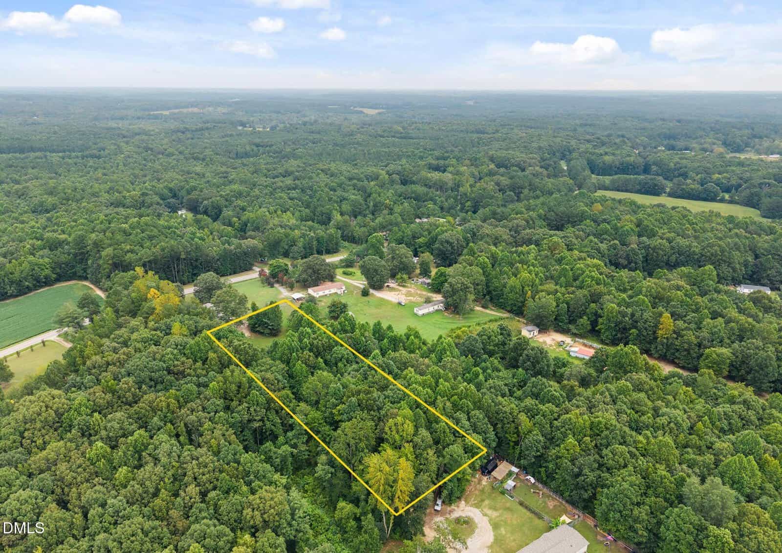 Aerial view of a wooded rural area with a rectangular plot of land outlined in yellow, surrounded by dense trees, scattered homes, and open fields under a partly cloudy sky.