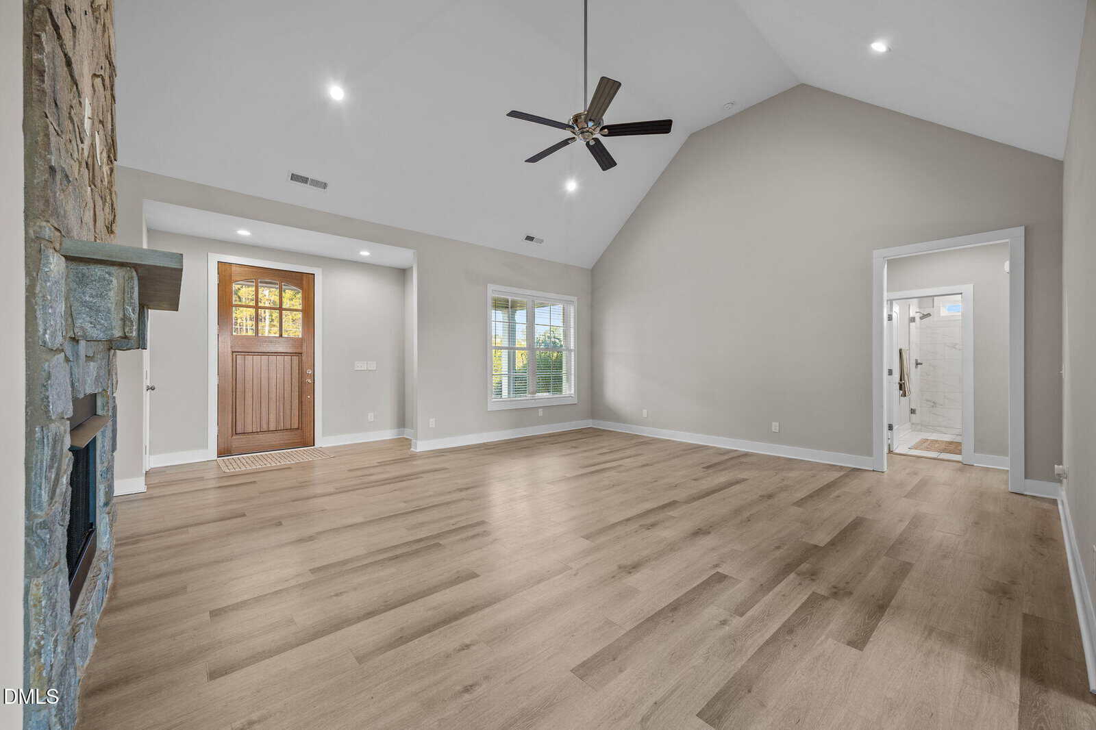 Spacious living room with vaulted ceiling, light wood floors, a stone fireplace, a large window, and a wooden front door; open doorway leads to a bathroom with white tiles.