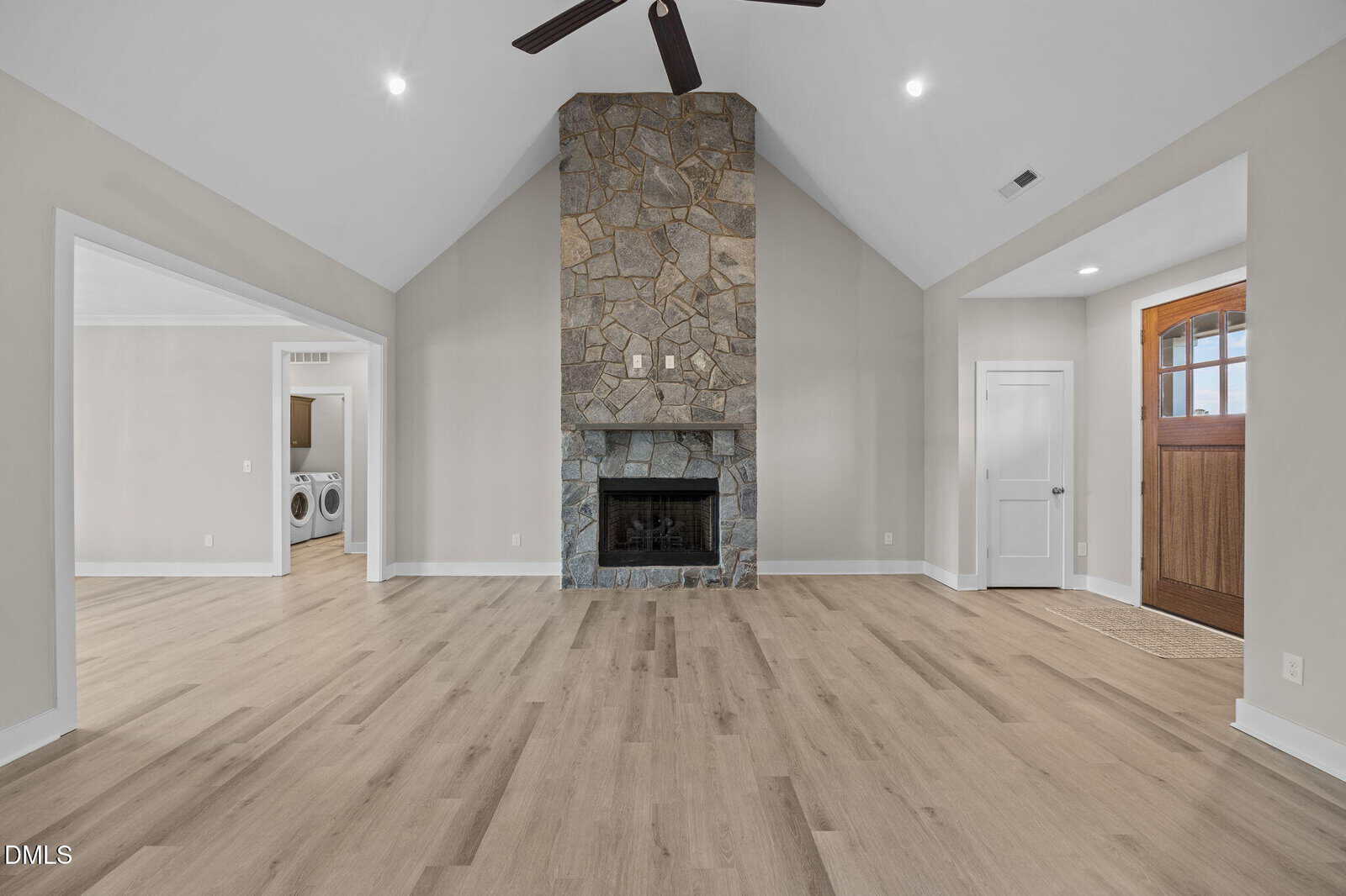 A spacious living room with light wood floors, a high vaulted ceiling, a central stone fireplace, and neutral walls. Visible are a laundry room to the left and a wooden front door to the right.