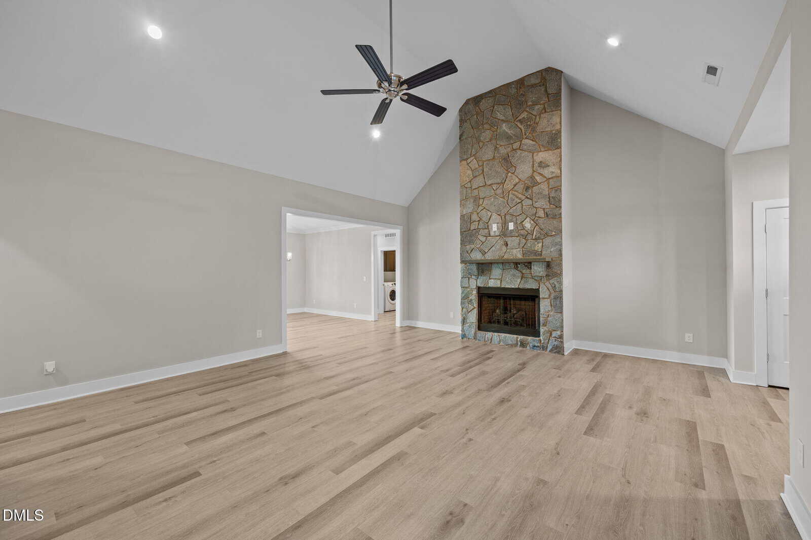 Spacious living room with high vaulted ceiling, light wood flooring, and a large stone fireplace; neutral-colored walls, modern recessed lighting, and a black ceiling fan. Doorways lead to adjacent rooms.
