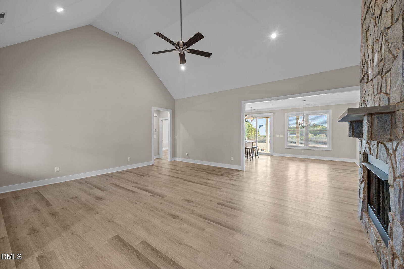 Spacious, empty living room with vaulted ceiling, light wood flooring, stone fireplace, neutral walls, ceiling fan, and large windows letting in natural light from an adjoining dining area.