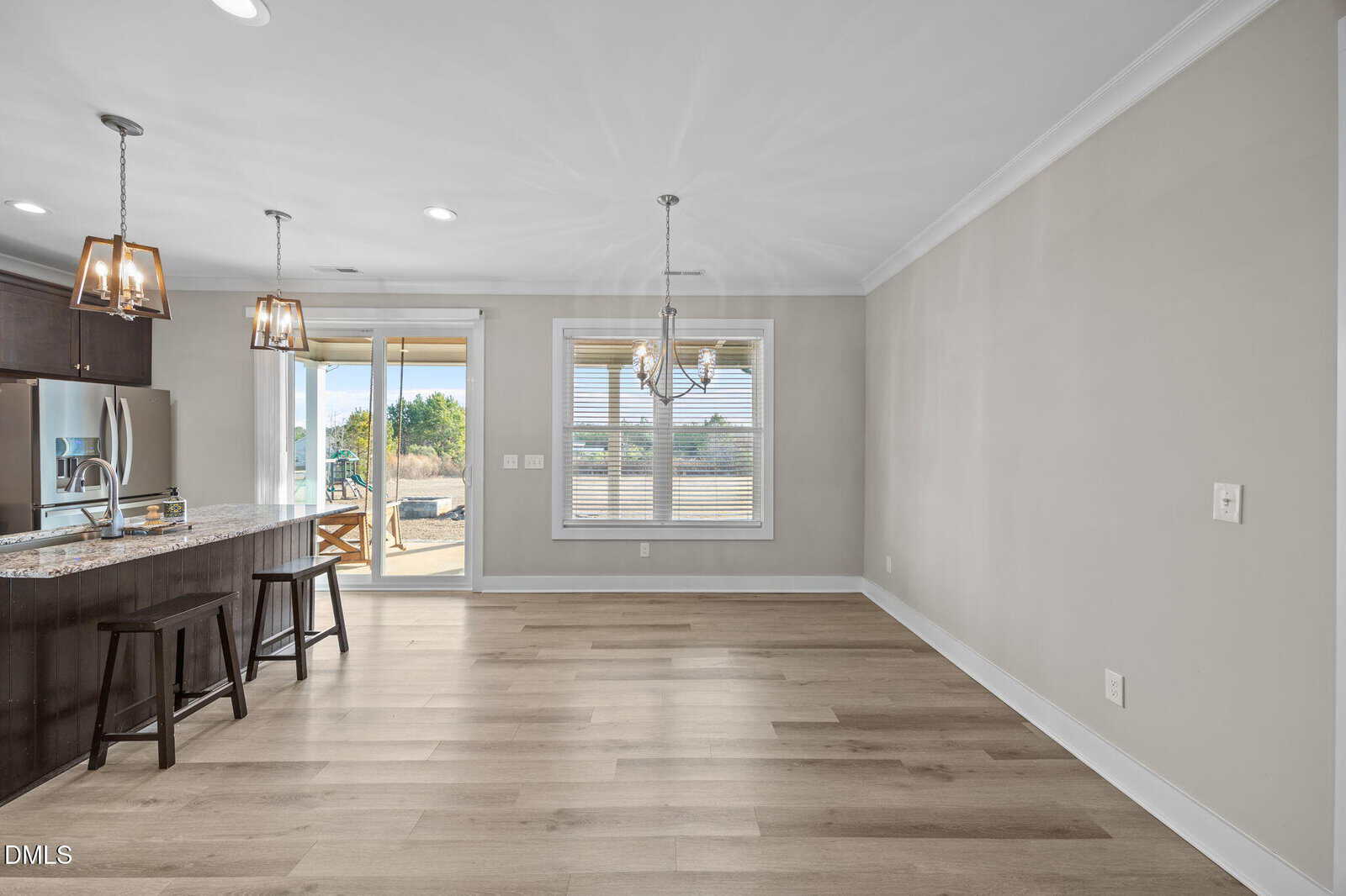 Modern kitchen and dining area with wooden floors, pendant lighting, stainless steel appliances, and large windows; sliding glass door opens to an outdoor patio with a view of greenery.