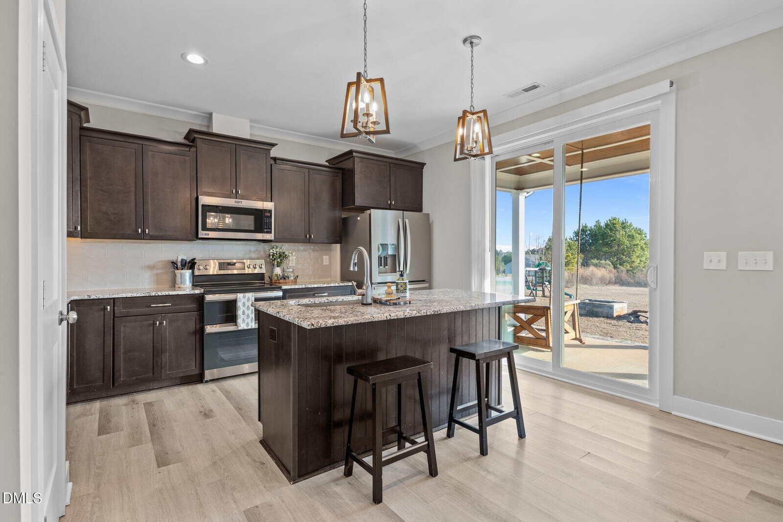 Modern kitchen with dark wood cabinets, stainless steel appliances, an island with granite countertop, two black stools, hanging pendant lights, and sliding glass doors leading to an outdoor patio with a scenic view.