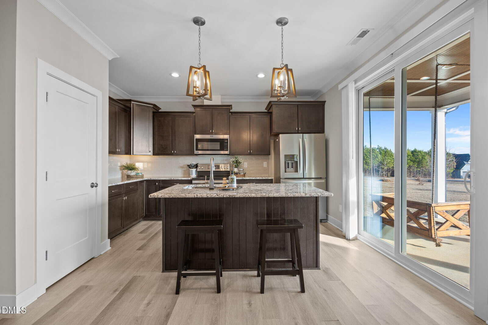 Modern kitchen with dark wood cabinets, a granite island with two stools, stainless steel appliances, two pendant lights, and sliding glass doors leading to a patio with outdoor seating and scenic mountain views.