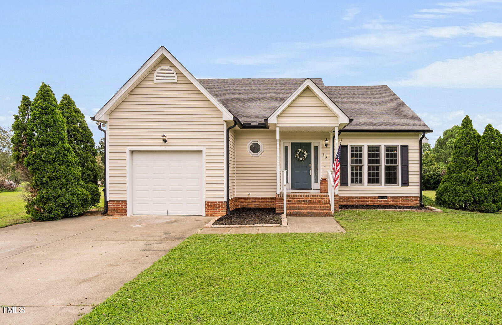 Charming single-story beige home at 65 Linnett Court, Youngsville, with a one-car garage, front porch with steps, American flag, and decorative wreath. Enjoy a green lawn and tall shrubs under a partly cloudy sky.