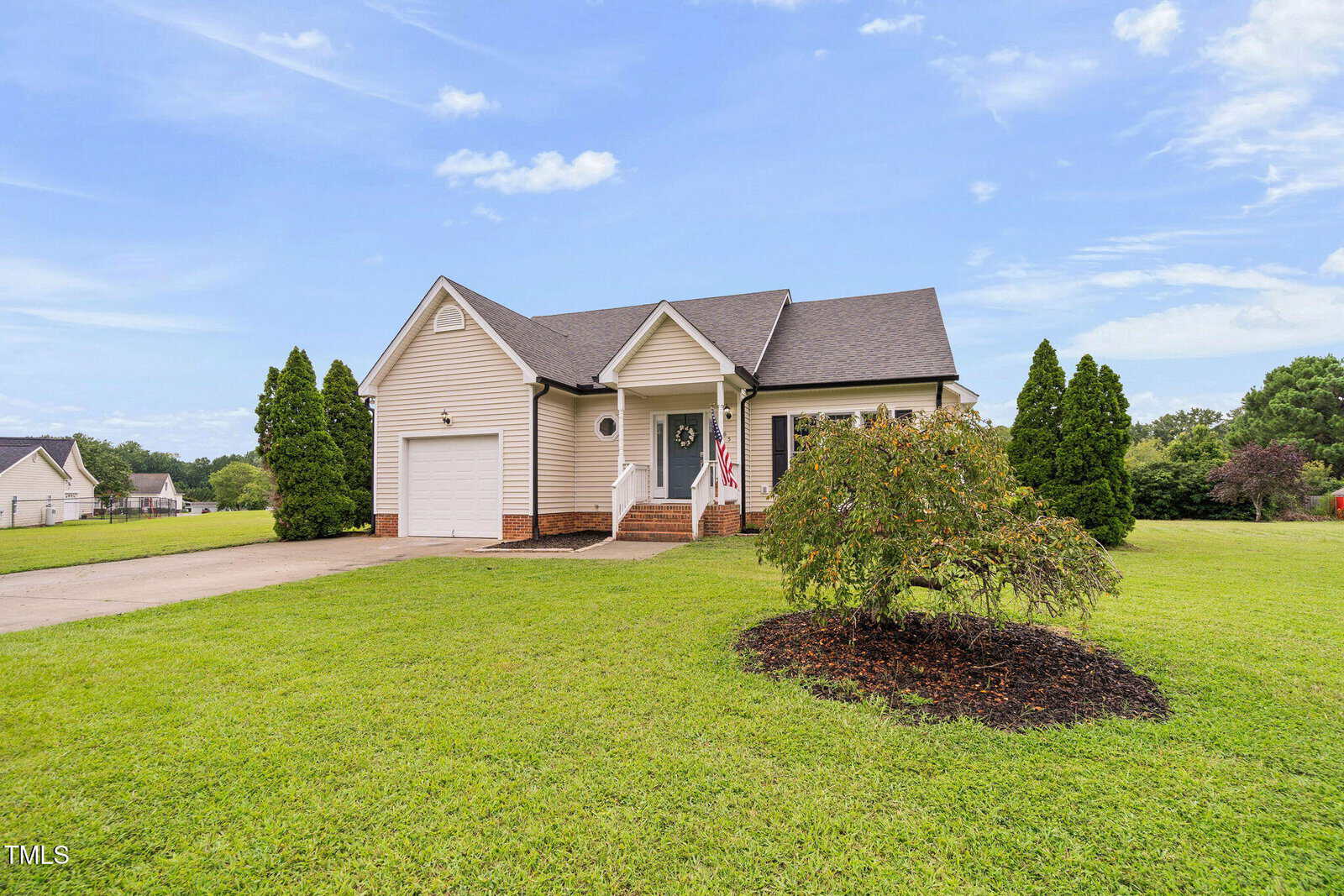 Single-story beige house with a gabled roof, front porch, American flag, and attached garage. The house sits on a large, neatly maintained lawn with a small tree and driveway under a partly cloudy sky.