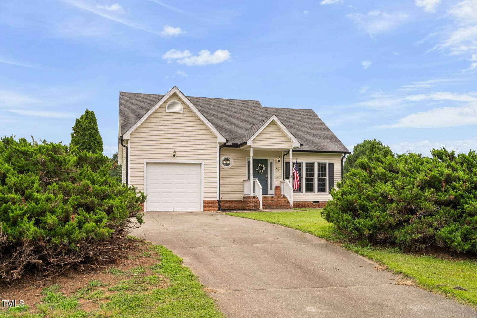 Single-story beige house with a front porch, attached garage, and American flag. Curved driveway leads to the garage, bordered by trimmed bushes, under a blue sky with scattered clouds.
