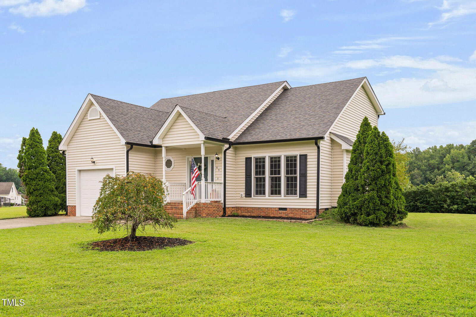 Single-story beige house with dark roof, attached garage, front porch with white railing, American flag, large windows, and well-kept lawn, surrounded by bushes and trees under a blue sky.