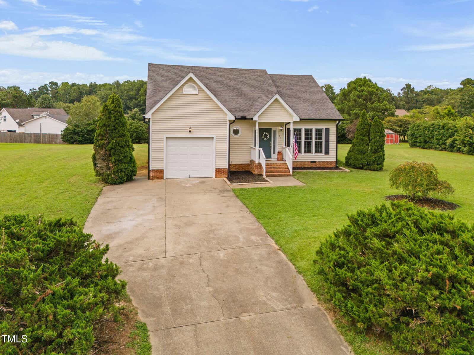 Single-story beige house with a front porch, attached garage, and American flag. The home is surrounded by a large green lawn, shrubs, and trees, with a concrete driveway leading to the garage.