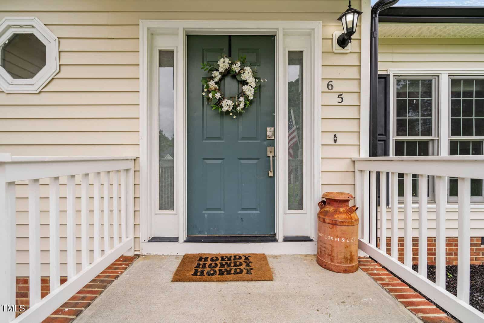 A teal front door with a floral wreath, flanked by sidelights, white railings, and a doormat that says “HOWDY” three times. An old milk can sits to the right of the door on a brick porch.