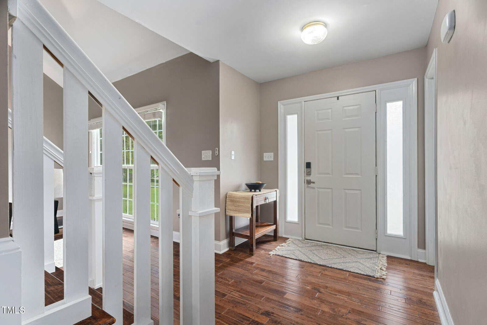 A bright entryway with a white front door, sidelights, a small rug, a wooden console table with a bowl, and hardwood floors. White stairs with wooden tops are visible on the left.