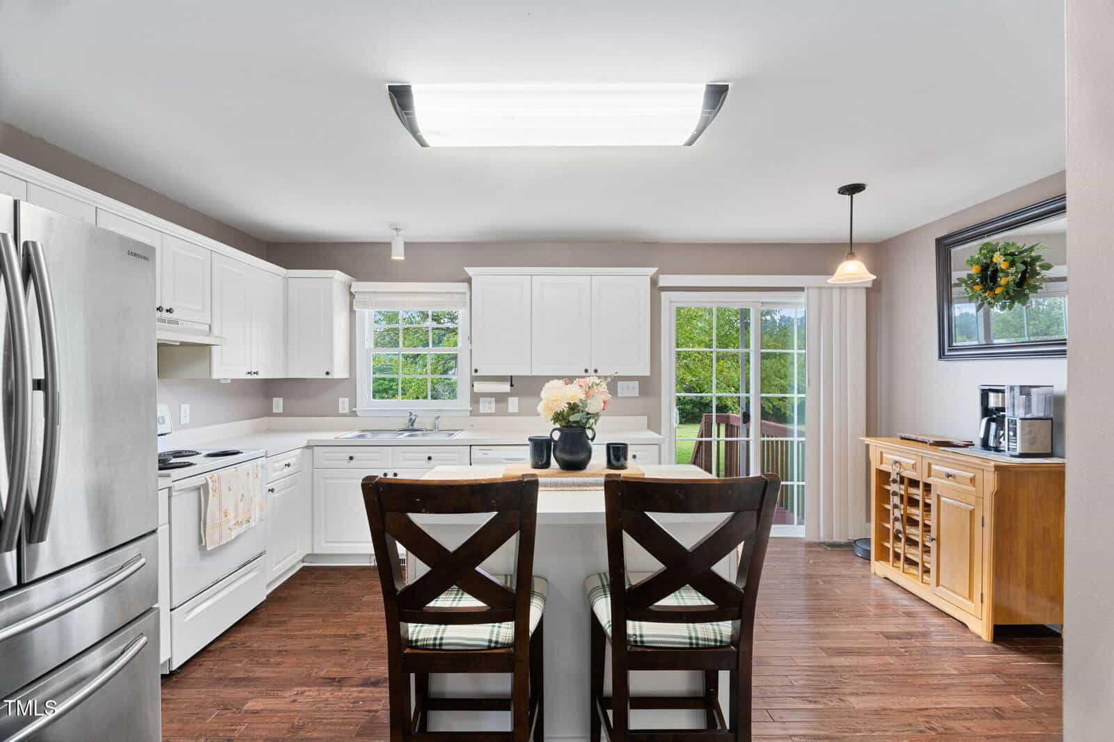 Bright, modern kitchen with white cabinets, stainless steel appliances, a central island with two wooden chairs, wood flooring, and large windows letting in natural light. A coffee station and wine rack are on the right.