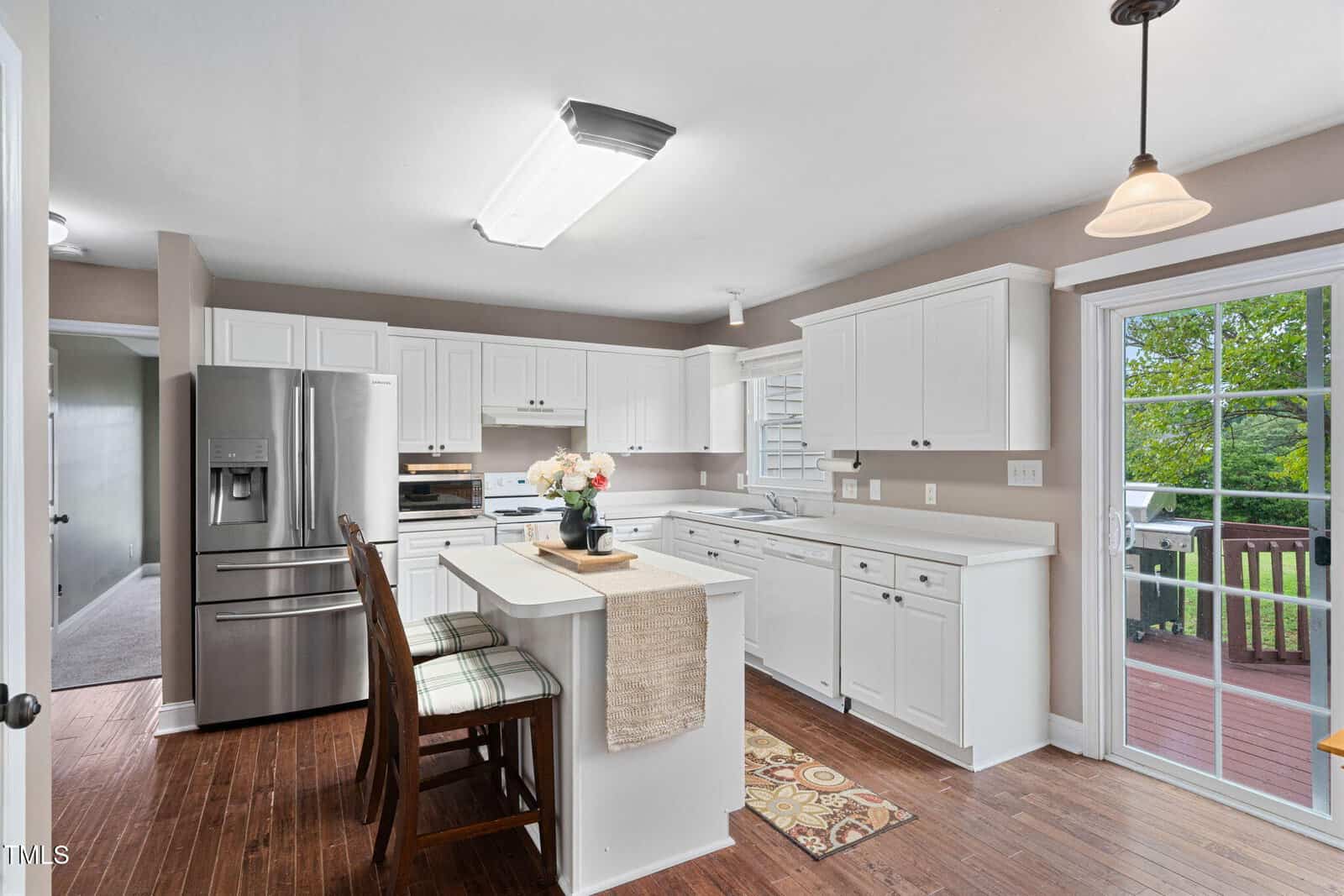 Bright kitchen with white cabinets, stainless steel appliances, an island with two chairs, wood floors, and a glass door leading to a deck with greenery visible outside. A pendant light hangs above the island.