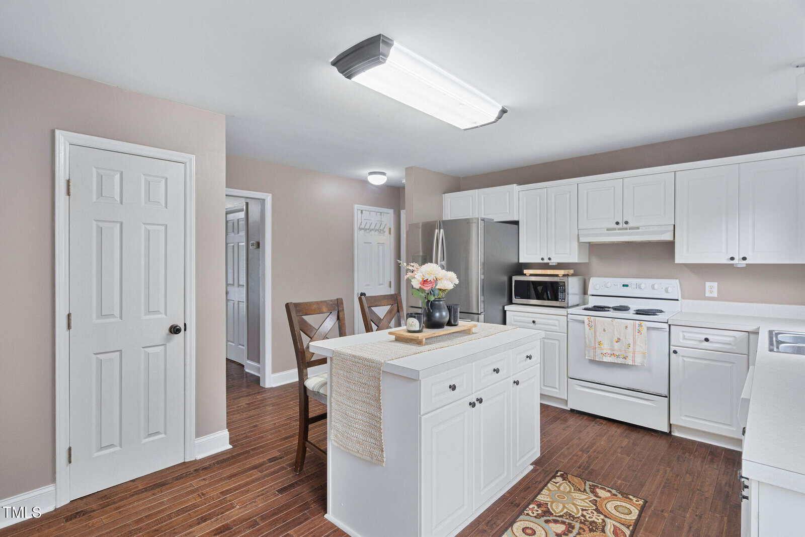 Bright kitchen with white cabinets, island, and appliances. A floral centerpiece sits on the island, which has two chairs. Hardwood floors and beige walls give a warm feel. Doorways and hall visible in the background.