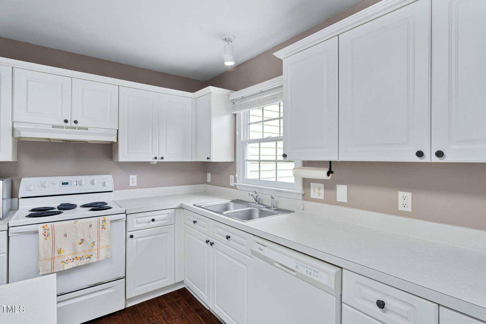 Bright kitchen with white cabinets, countertops, and appliances, including a stove, dishwasher, and double sink under a window with natural light. Brown walls and dark wood flooring complete the space.