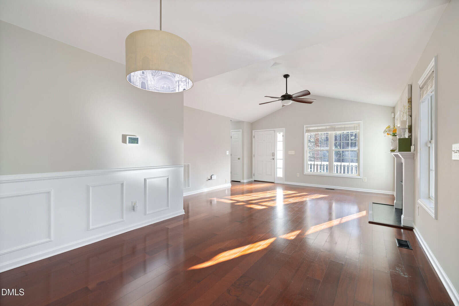 Bright, empty living room with polished wood floors, large window, white walls, wainscoting, ceiling fan, hanging light fixture, and a white front door letting in natural sunlight.