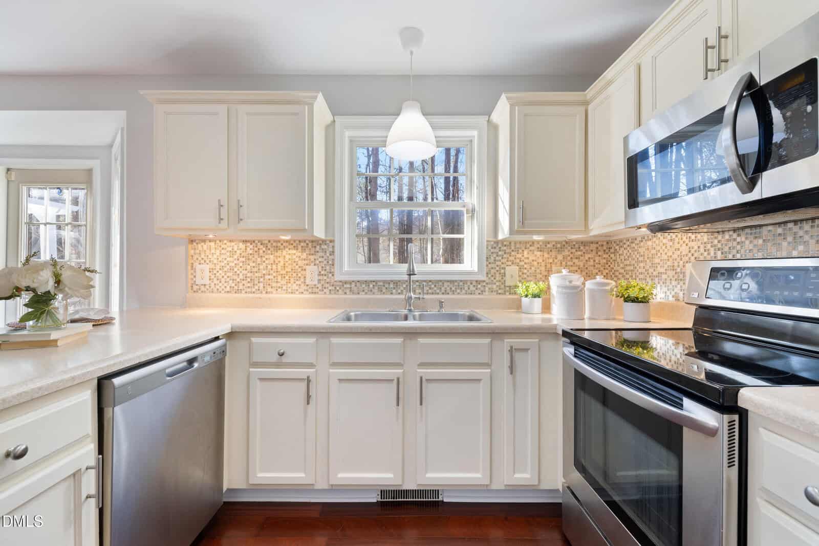 Bright, modern kitchen with white cabinets, stainless steel appliances, a mosaic tile backsplash, a double sink under a window, and potted plants on the countertop. Sunlight streams in through the window.