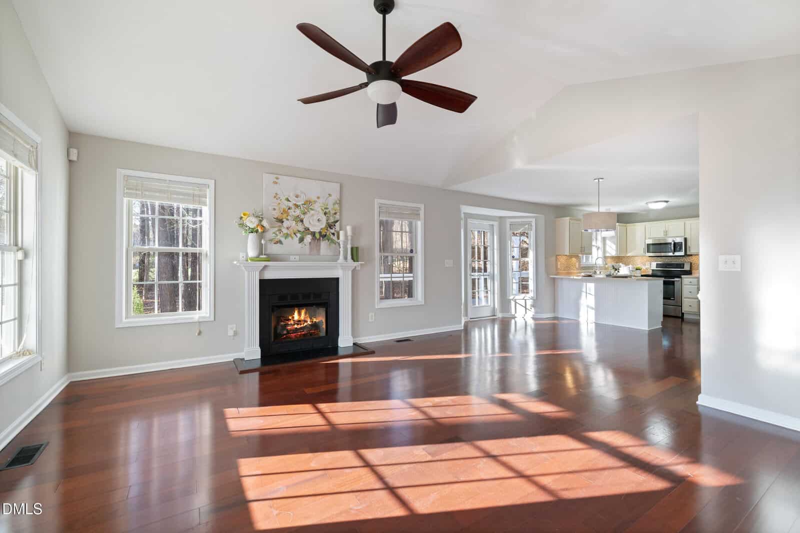 Bright, spacious living room with hardwood floors, large windows, a ceiling fan, a fireplace with floral artwork above, and an open view into a modern kitchen with white cabinets and stainless steel appliances.