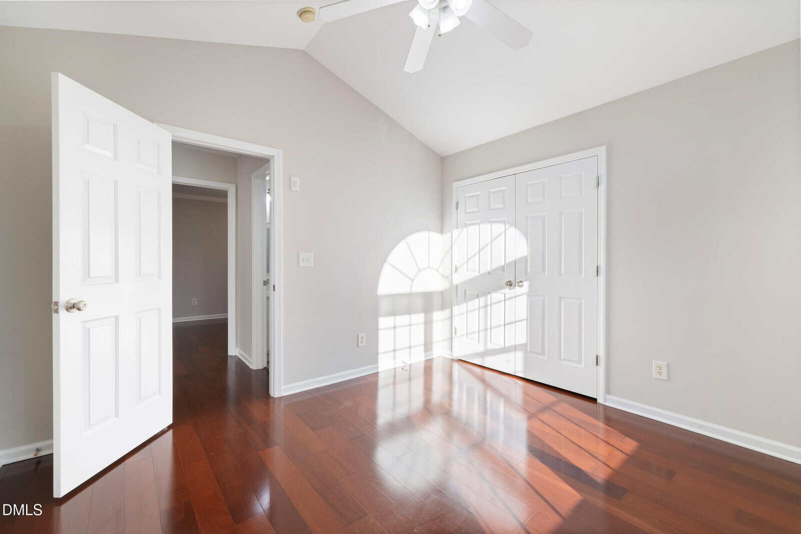 Bright, empty room with hardwood floors, white walls, and two white double doors. Sunlight streams through a semicircular window, casting shadows on the floor. A ceiling fan is visible above.