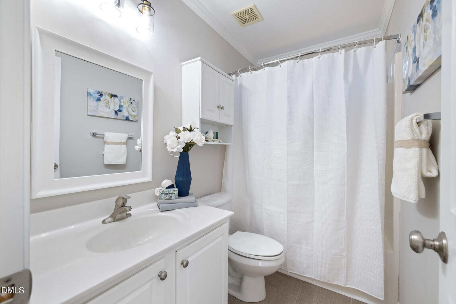 A modern bathroom with white cabinetry, a sink, toilet, shower with a white curtain, towel racks, a wall cabinet, floral decor, and a blue vase with white flowers on the toilet tank.