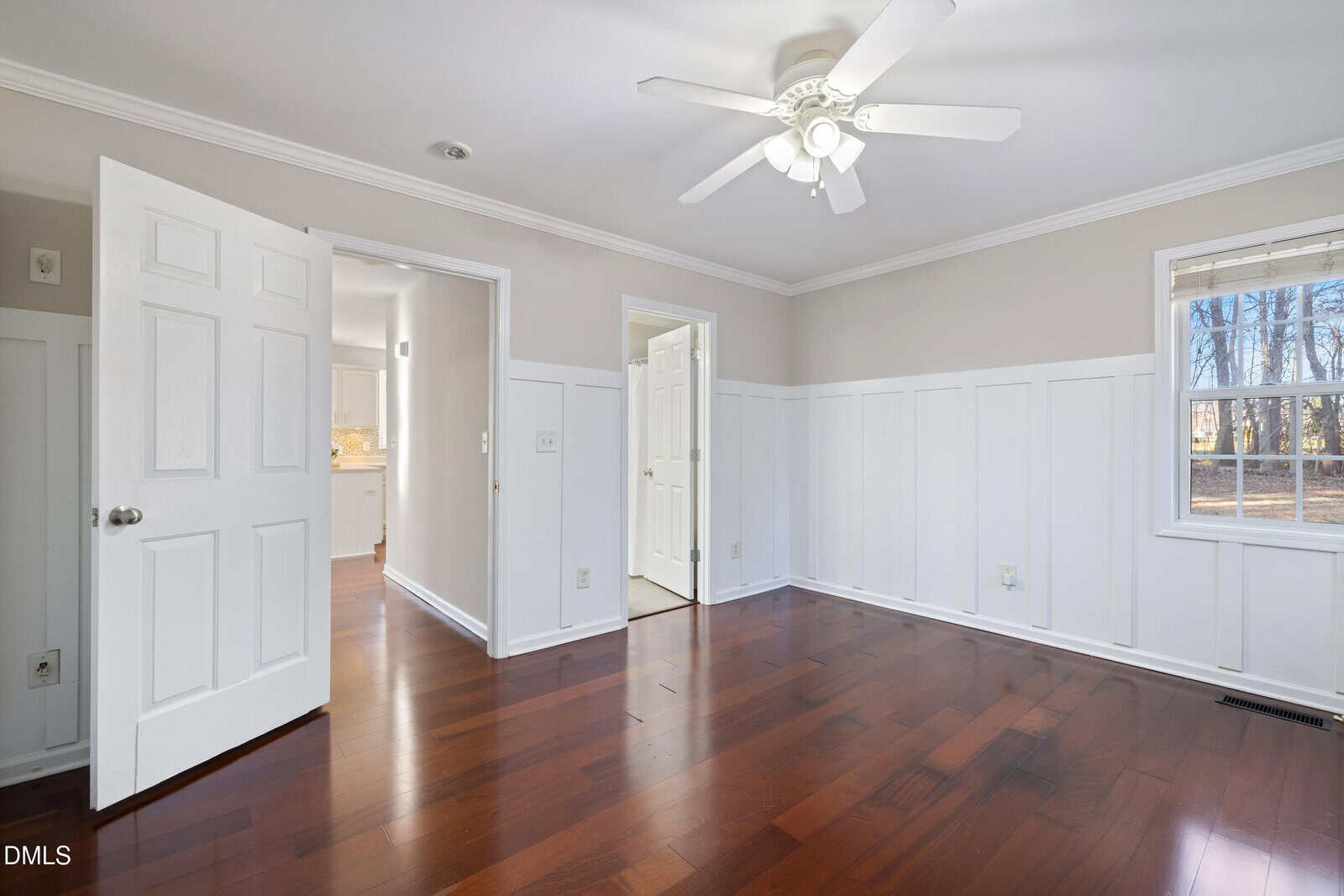 A bright, empty room with white wainscoting, light beige walls, dark hardwood floors, a ceiling fan, an open door leading to another room, and a window with a view of trees outside.
