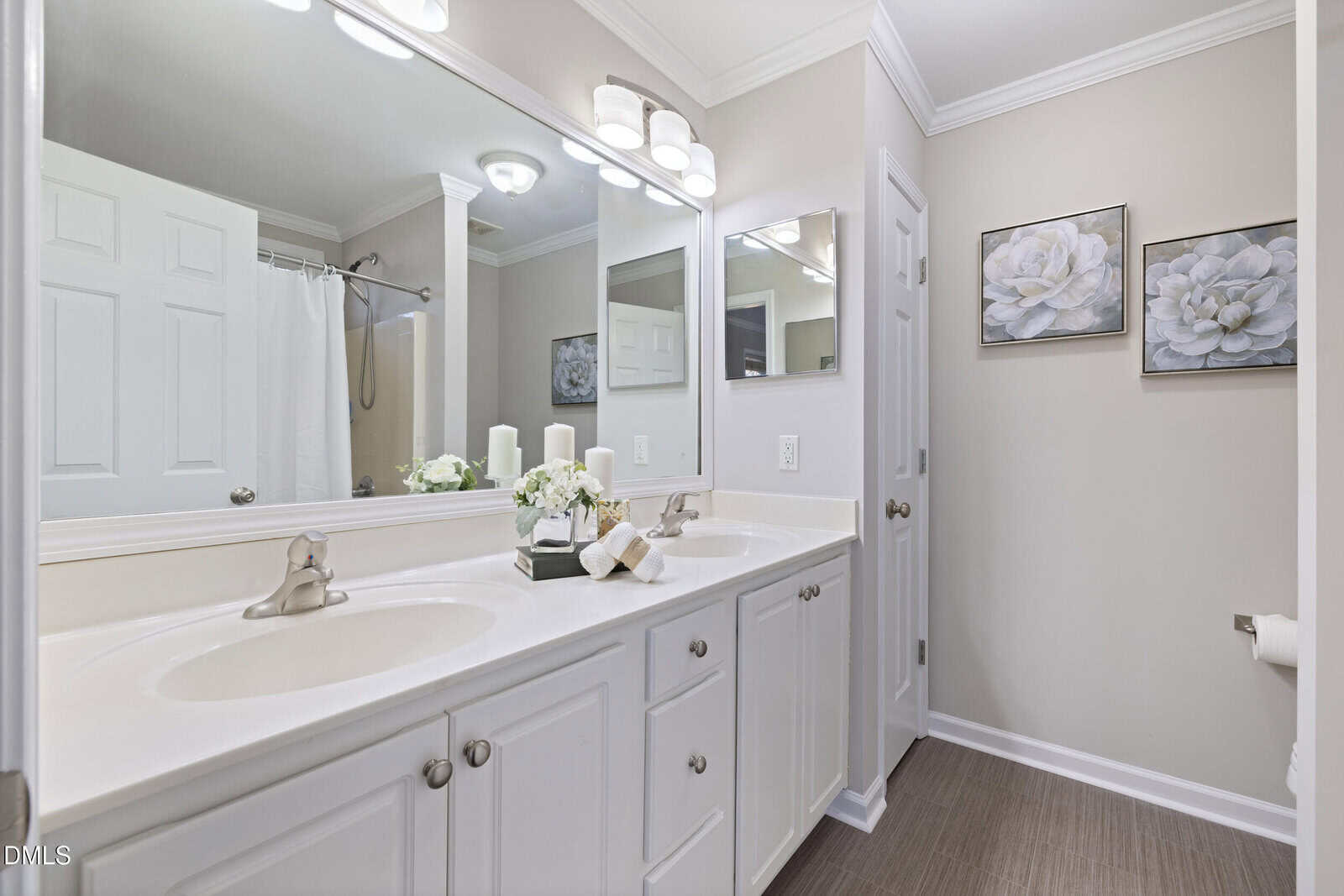 Bright, modern bathroom with a double-sink vanity, large mirror, white cabinets, neutral walls, and floral artwork. Towels, candles, and a small plant are neatly arranged on the counter. A shower curtain is visible in the background.