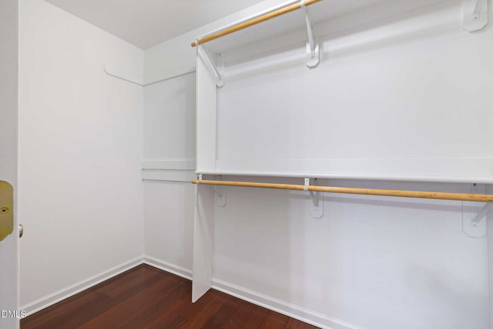 Empty walk-in closet with white walls, wooden rods for hanging clothes, white shelves, and dark wood flooring.