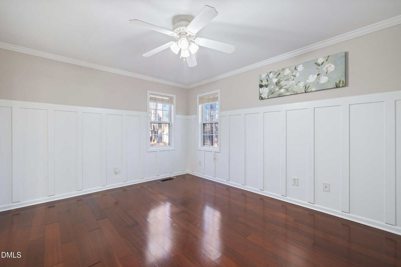 Bright empty room with white paneled walls, dark hardwood floors, two windows, a ceiling fan with lights, and a floral painting above the wainscoting.