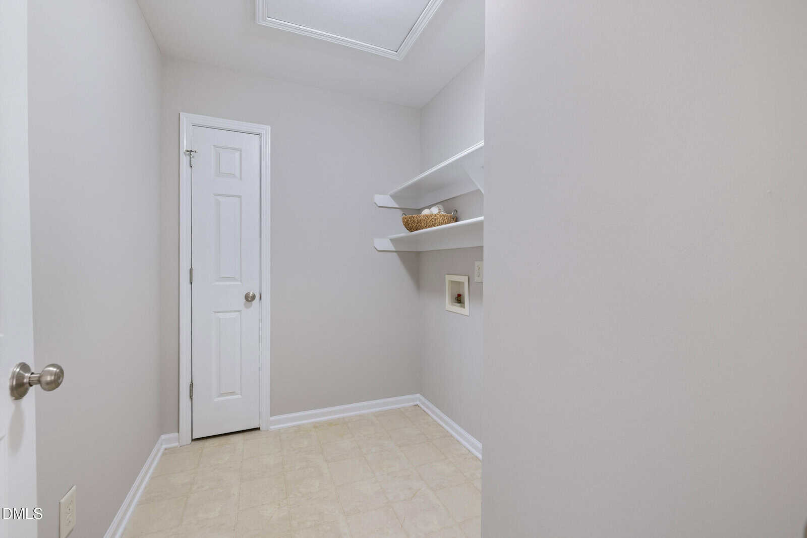Small, empty room with light-colored walls and floor, a closed white door, two white shelves holding a basket, and a small wall niche. Ceiling light is visible above.